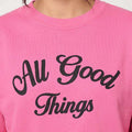 Candy pink sweatshirt close-up with black 'All Good Things' script print.
